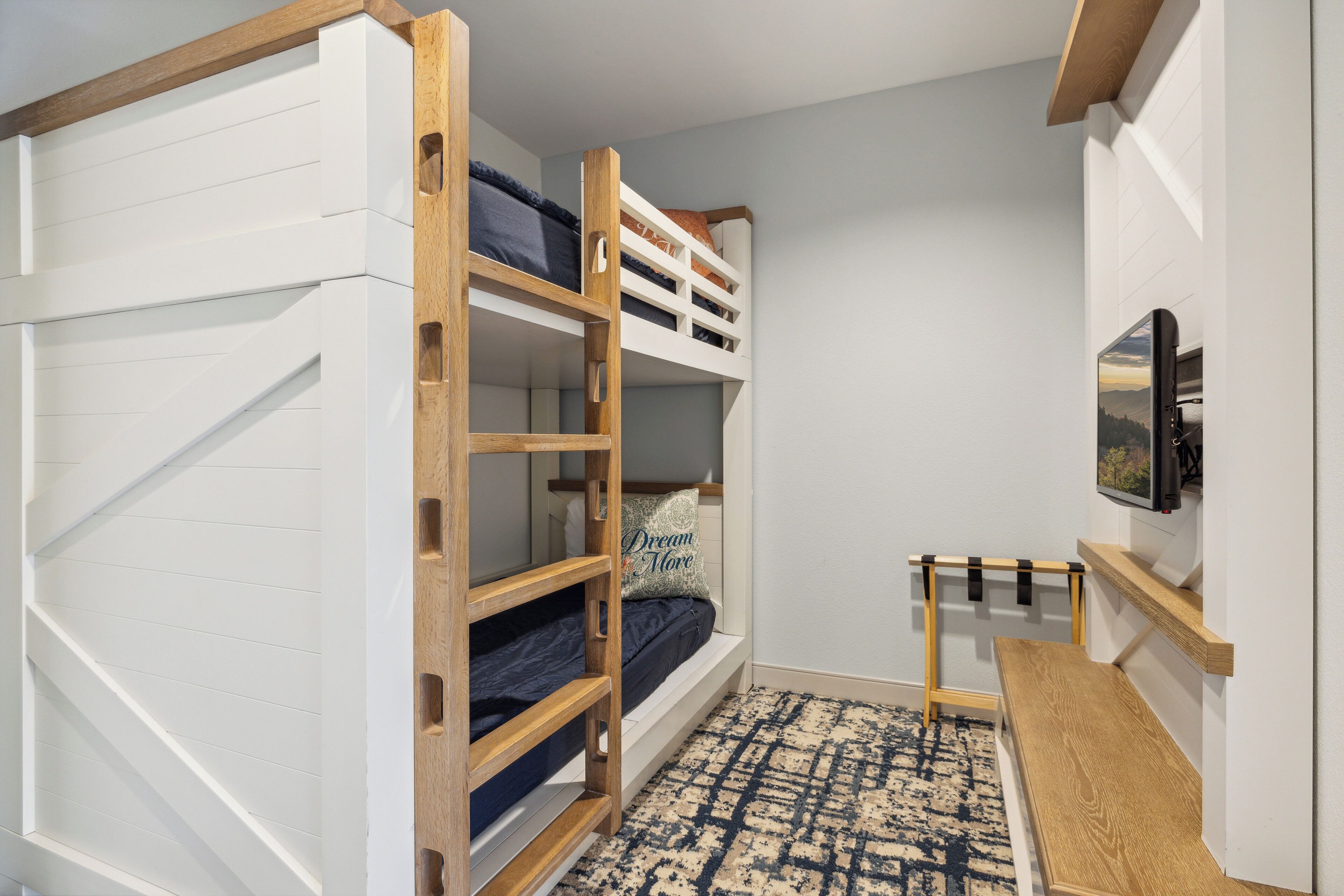 A bunk bed room with a TV on the wall and a wooden bench.