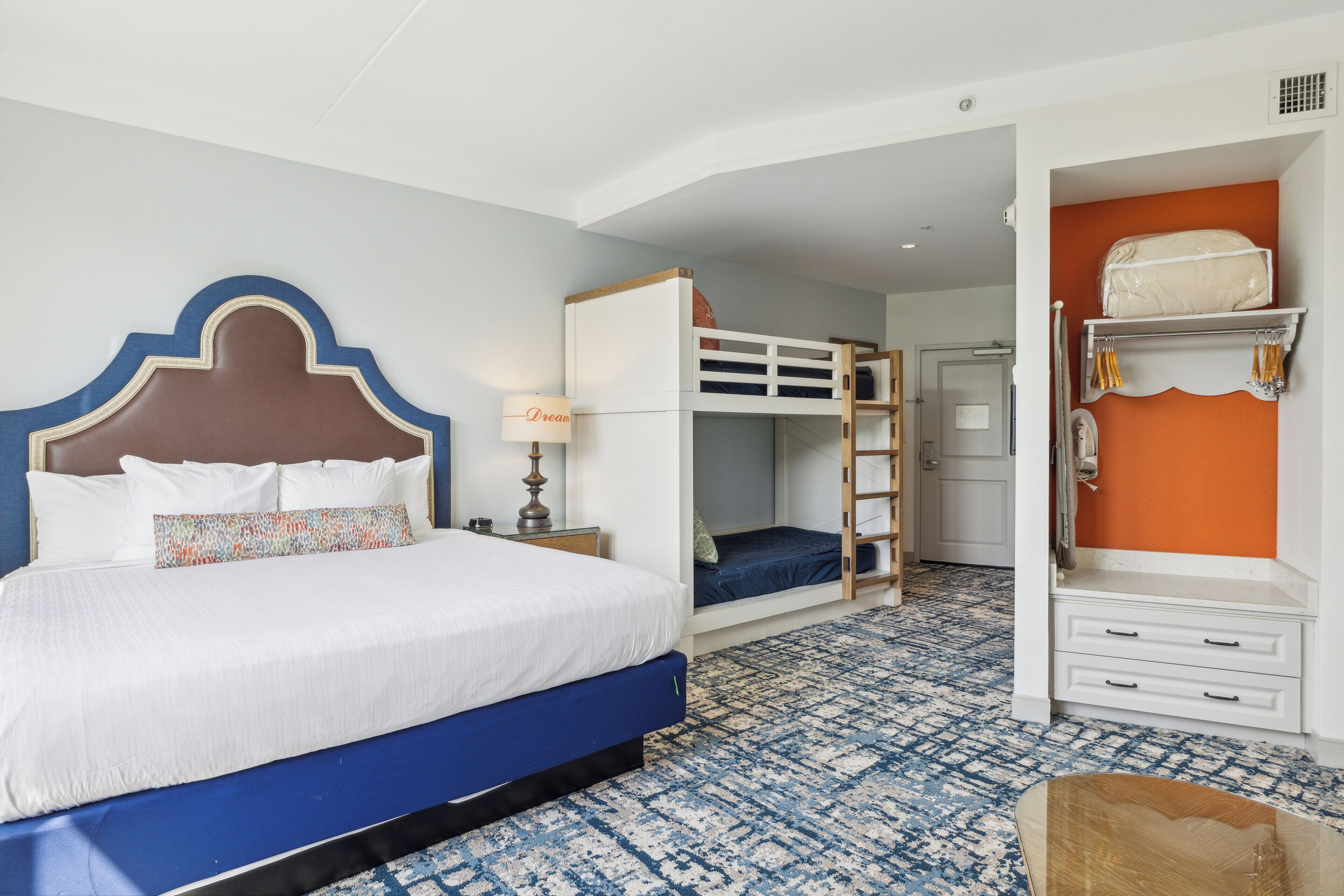 A hotel room with a large bed, a bunk bed, a desk with drawers, and a patterned carpet.
