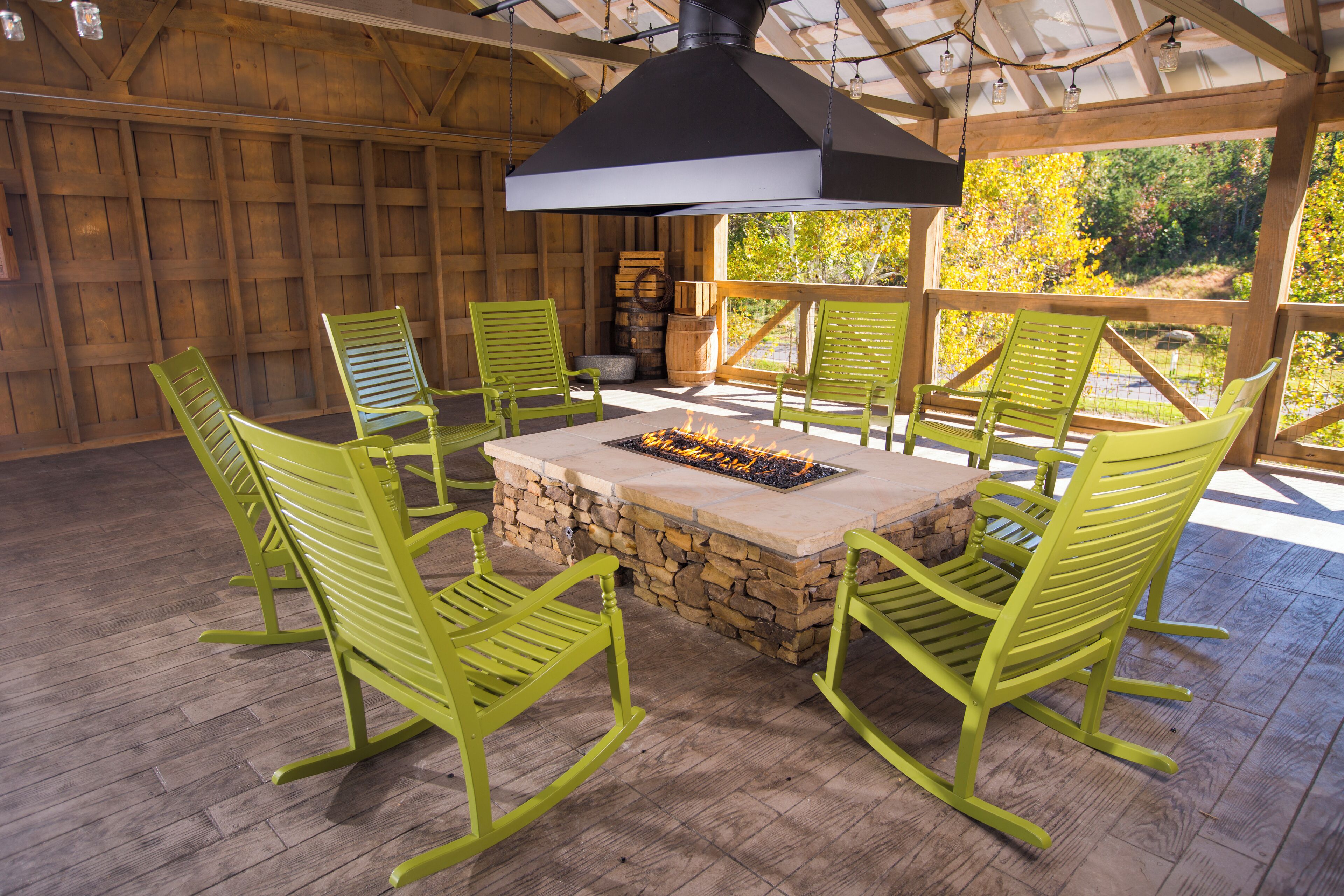 A wooden deck with a stone fire pit surrounded by green chairs, under a black chimney.