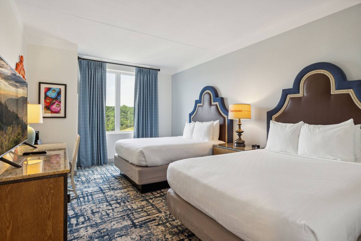 A hotel room with two beds, a desk with a lamp, a TV, and a window with curtains.