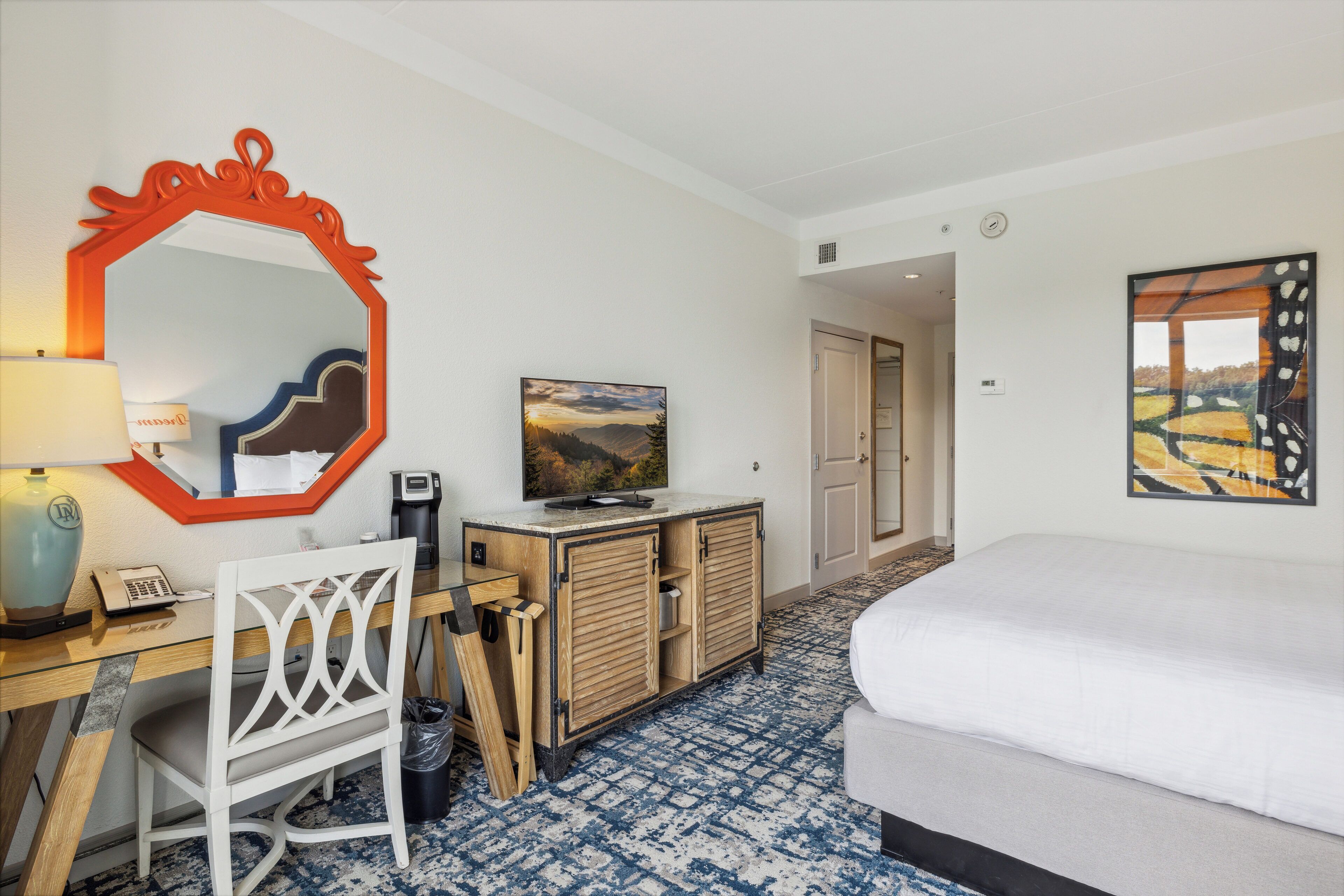 A hotel room with a bed, a desk with a chair, a mirror, a television, a cabinet, and a painting on the wall.