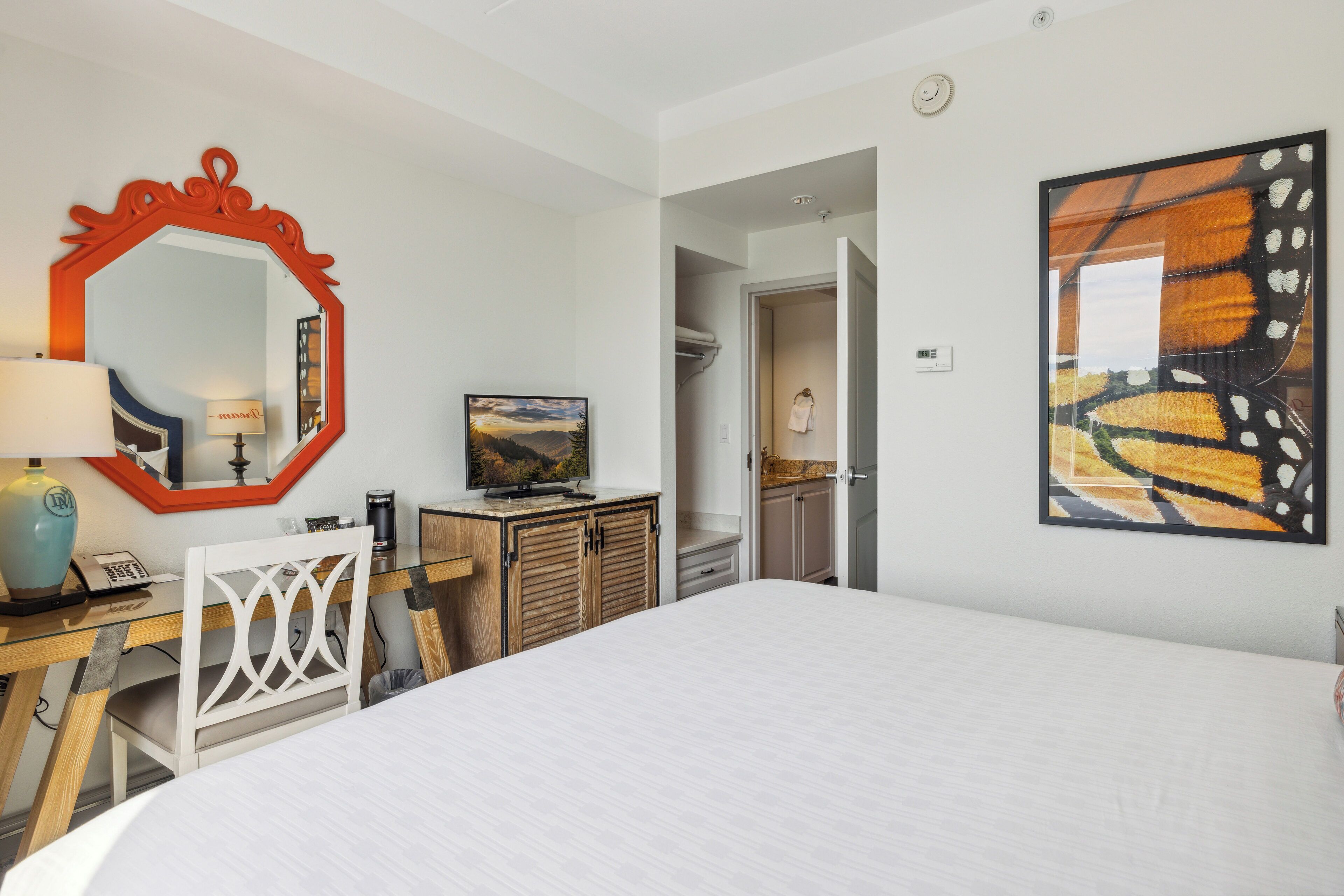 A hotel room with a large bed, a desk with a lamp, a television, and a mirror. There is a painting on the wall and a door leading to another room.