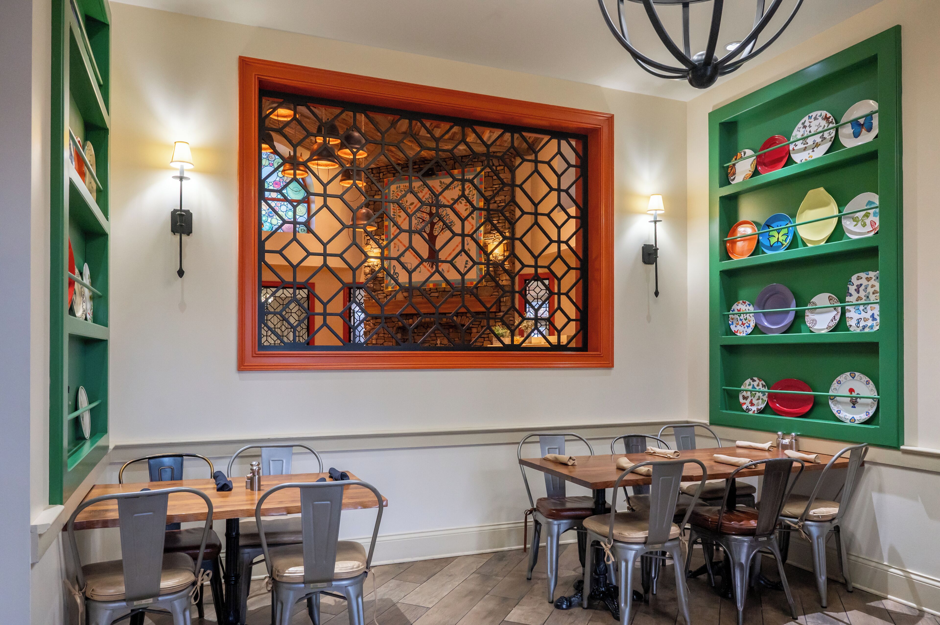 Un espace repas avec une table en bois et des chaises en métal, une fenêtre décorative ornée d’un motif géométrique et des luminaires fixés au mur.