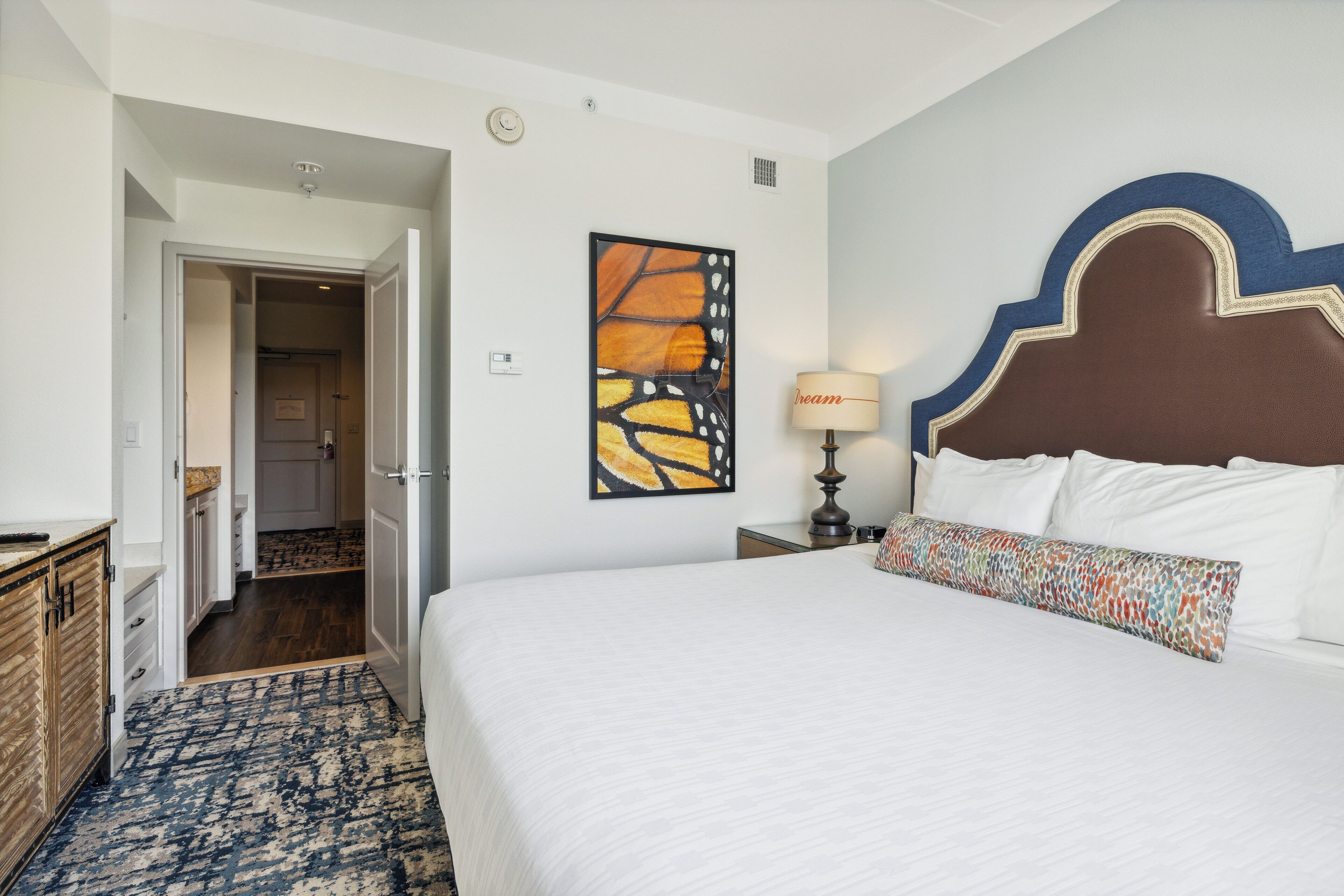 A hotel room with a large bed, a nightstand with a lamp, a wall clock, and a door leading to a hallway.