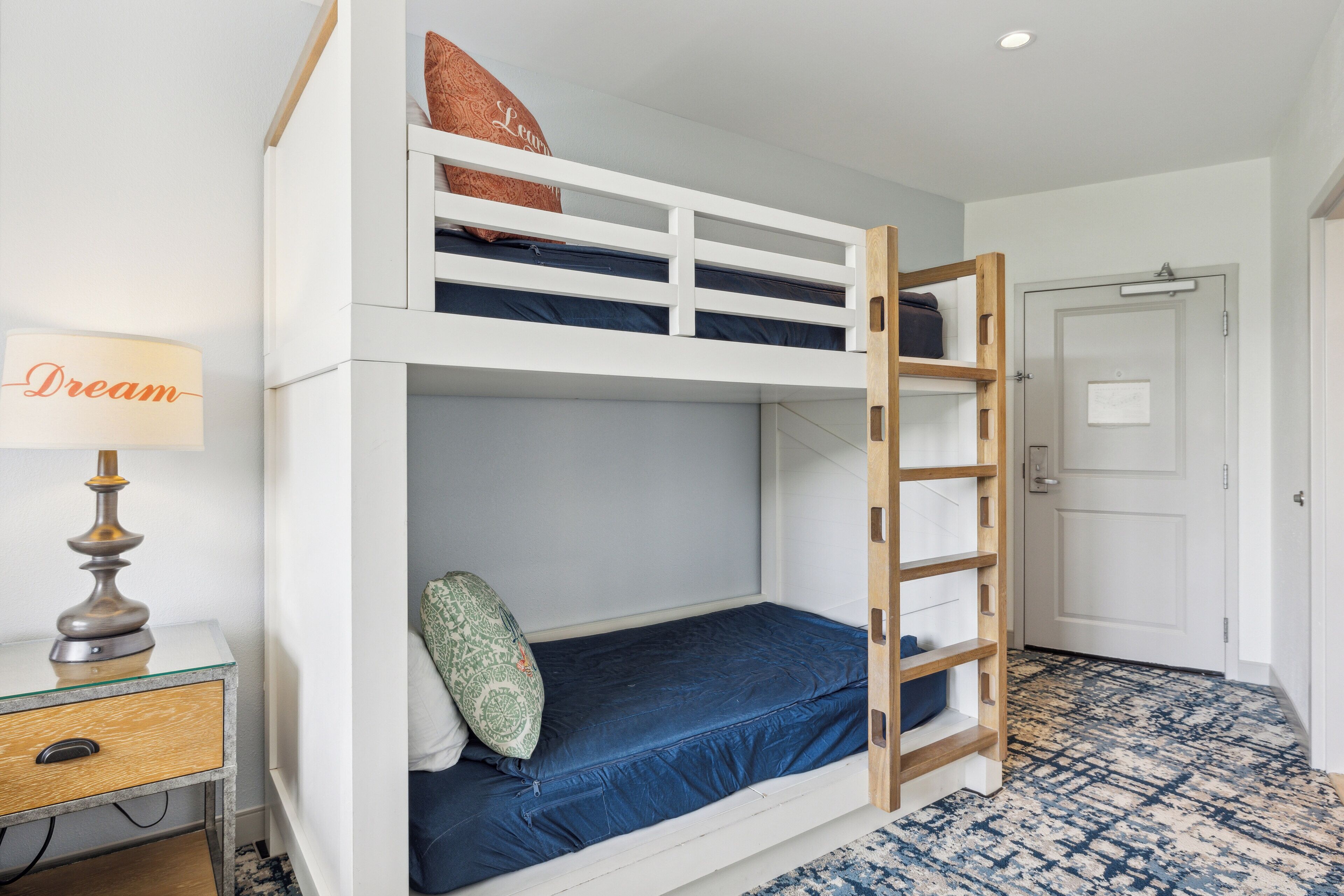 Una cama individual superpuesta con escalera, una mesita de noche con lámpara y una alfombra estampada.