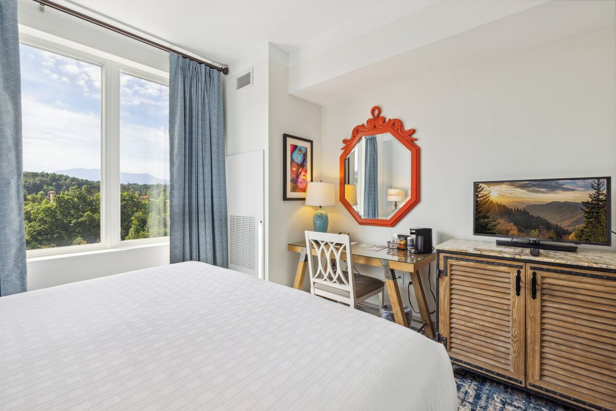 A hotel room with a large bed, a desk with a chair, a television, and a mirror.