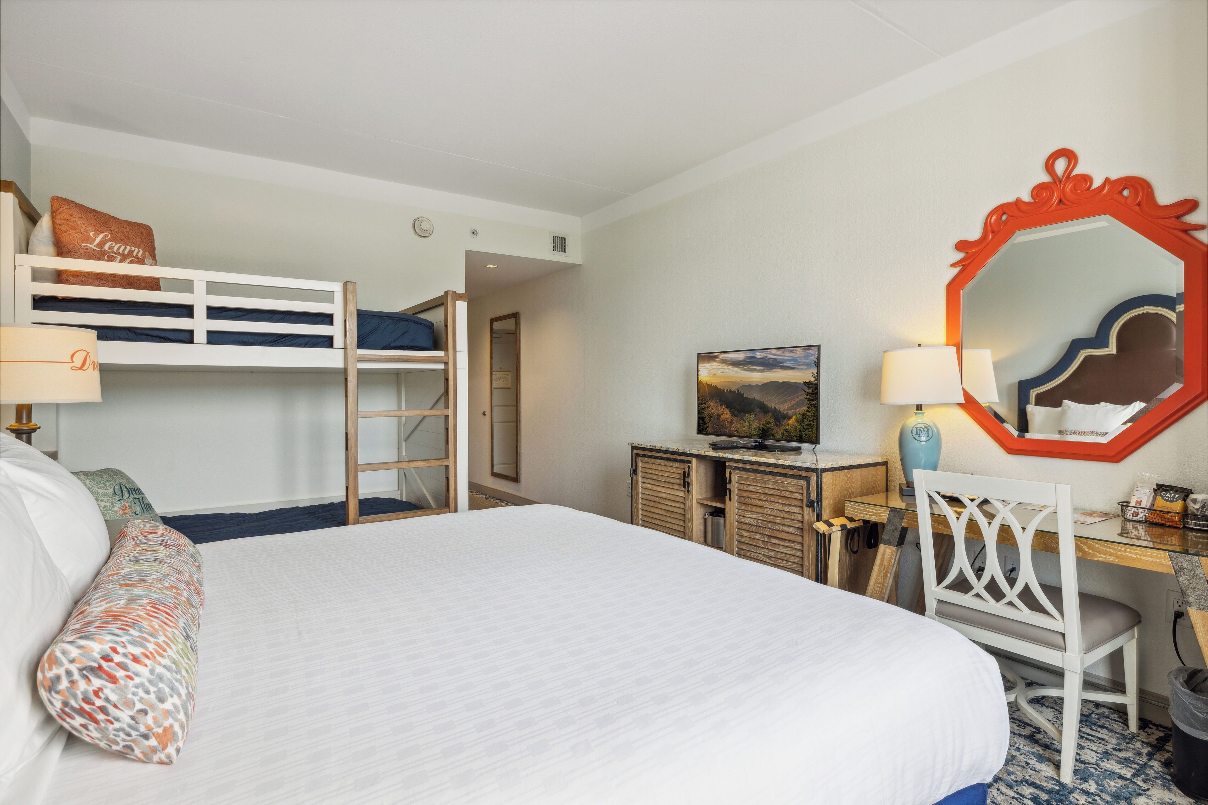 A hotel room with a bunk bed, a desk with a chair, a TV, and a large mirror.