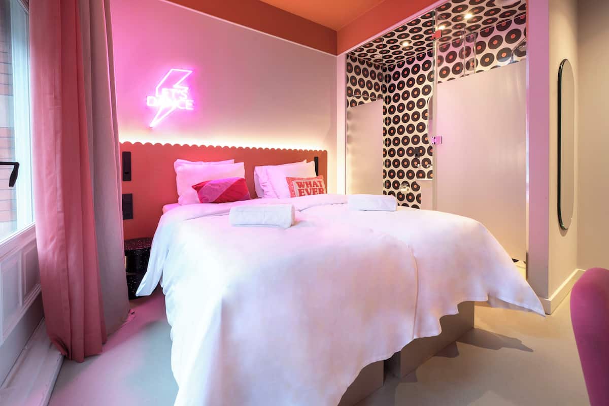 Un dormitorio moderno con una cama, un letrero de neón, papel tapiz estampado y una ventana con cortinas.