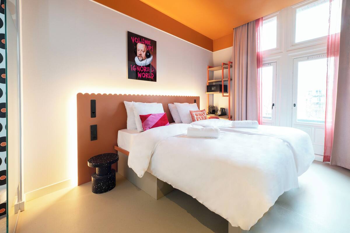 Habitación de hotel con una cama grande, un cartel en la pared, una mesita de noche y una ventana con cortinas.