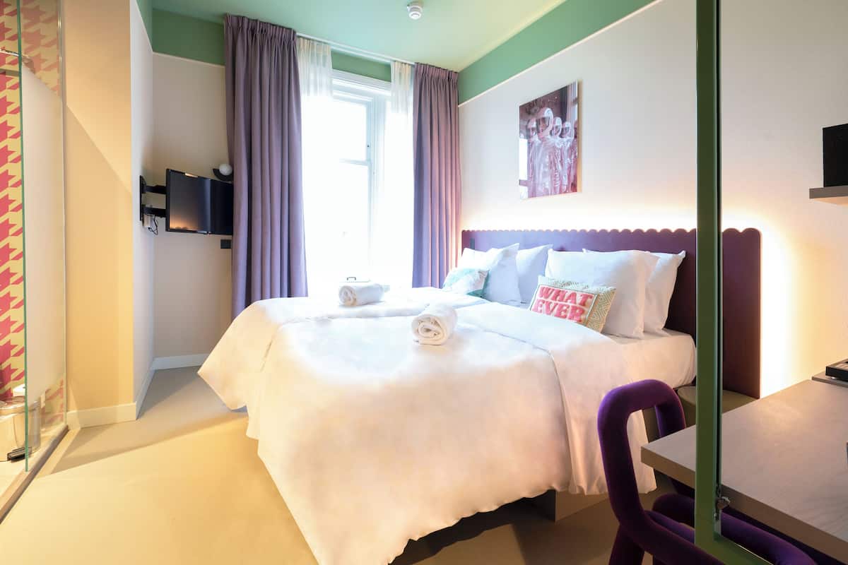 Una habitación de hotel con una cama, un televisor montado en la pared, una ventana con cortinas y una mesita con una silla.