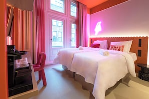 Habitación de hotel con dos camas, cafetera, una silla rosa y ventana con cortinas.