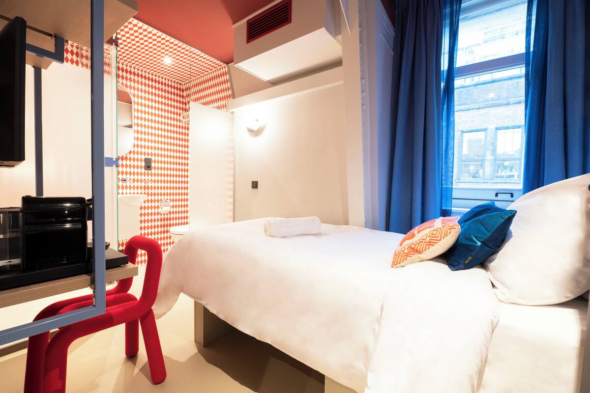Una habitación de hotel con cama, escritorio con cafetera, una silla roja y ventana con cortinas azules.