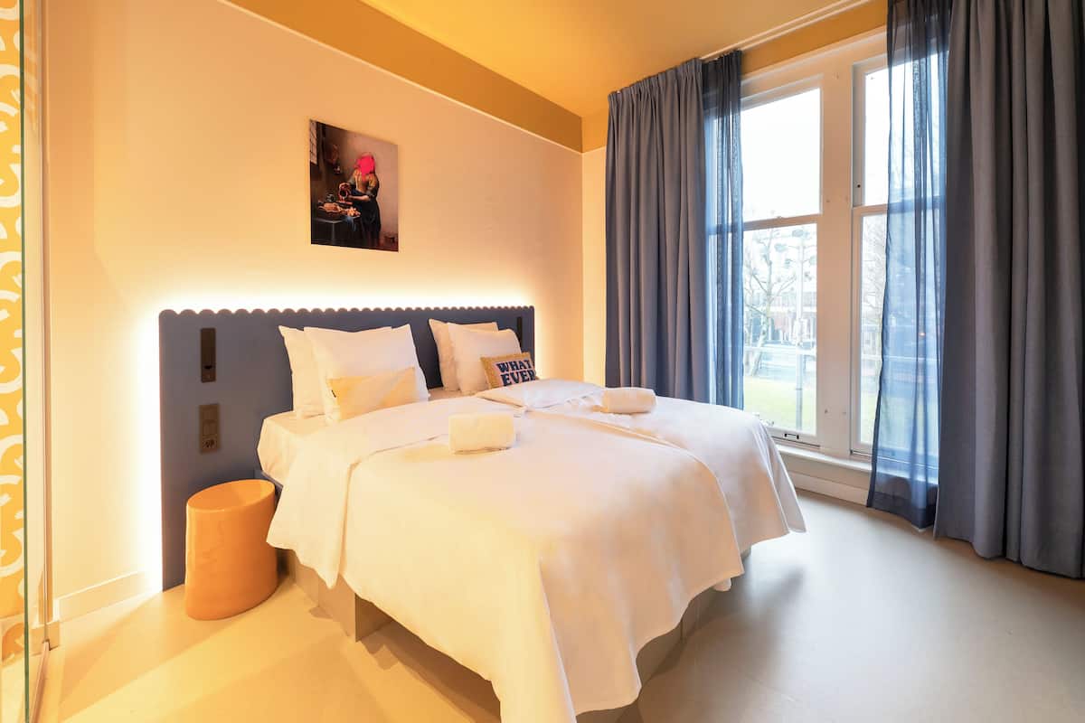 Habitación de hotel con una cama grande, una mesita de noche, un cubo de basura y una ventana con cortinas.