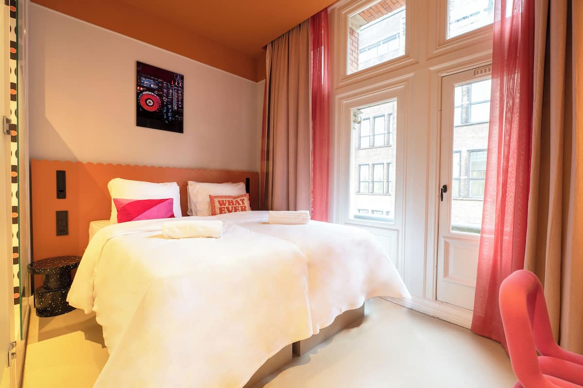 Habitación de hotel con una cama grande, una mesita, una silla y un ventanal con cortinas.