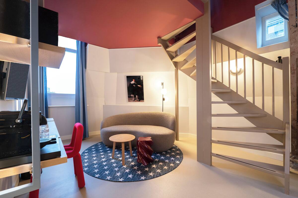 Una habitación moderna con un sofá curvo, una pequeña mesa redonda, una silla roja, un tapete con estampado de estrellas y una escalera con barandilla blanca.