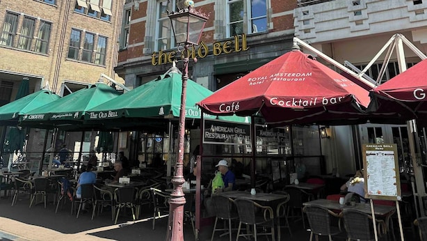 Un edificio con letreros que dicen 'HOTEL' en la fachada, mesas al aire libre con sombrillas y una farola.