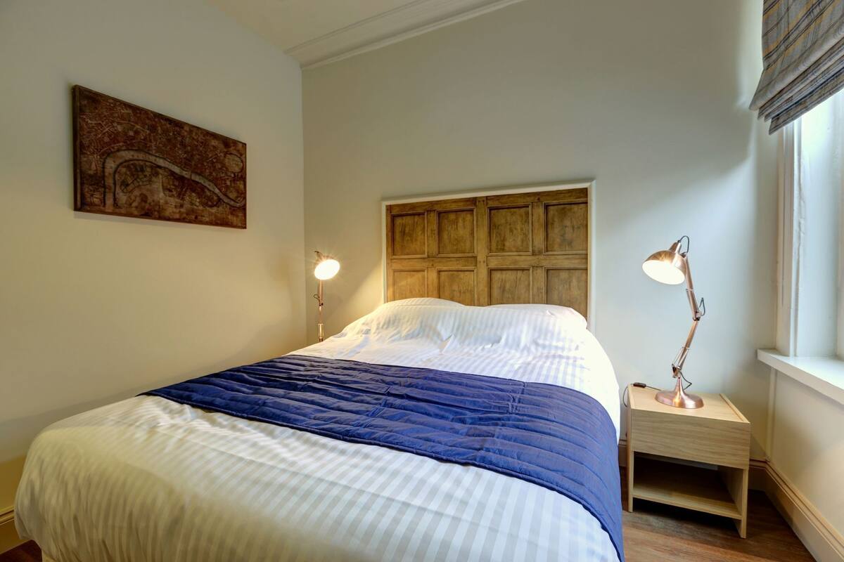 Ein Schlafzimmer mit einem Bett, einem Kopfteil, Nachttischlampen, einem Nachttisch, einem Fenster und einem Wandbild.