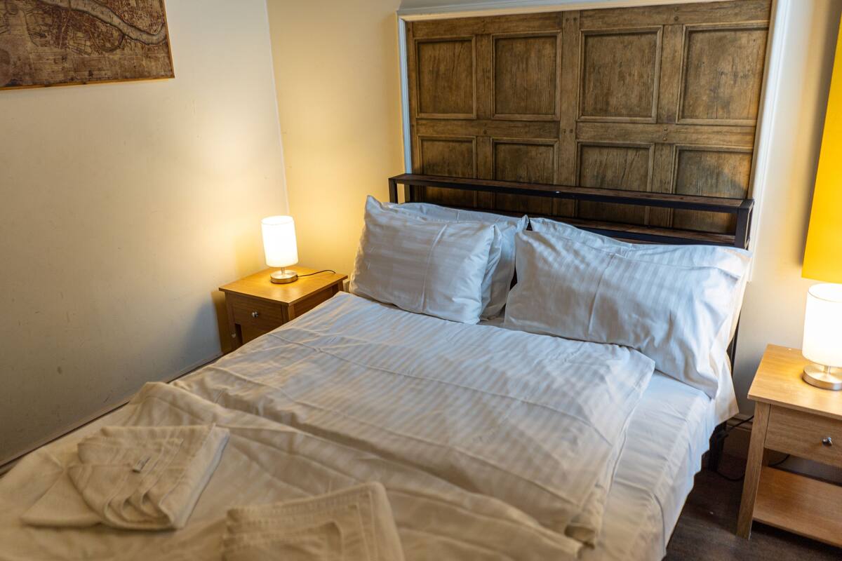 Ein ordentlich bezogenes Bett mit weißen Leinentüchern und einem hölzernen Kopfteil. Ein Nachttisch mit Lampe befindet sich links vom Bett.