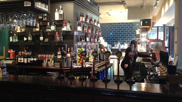 A bar area with shelves of liquor bottles, a bartender, and a counter with glasses and bottles. - Publove @ Exmouth Arms Euston (London)
