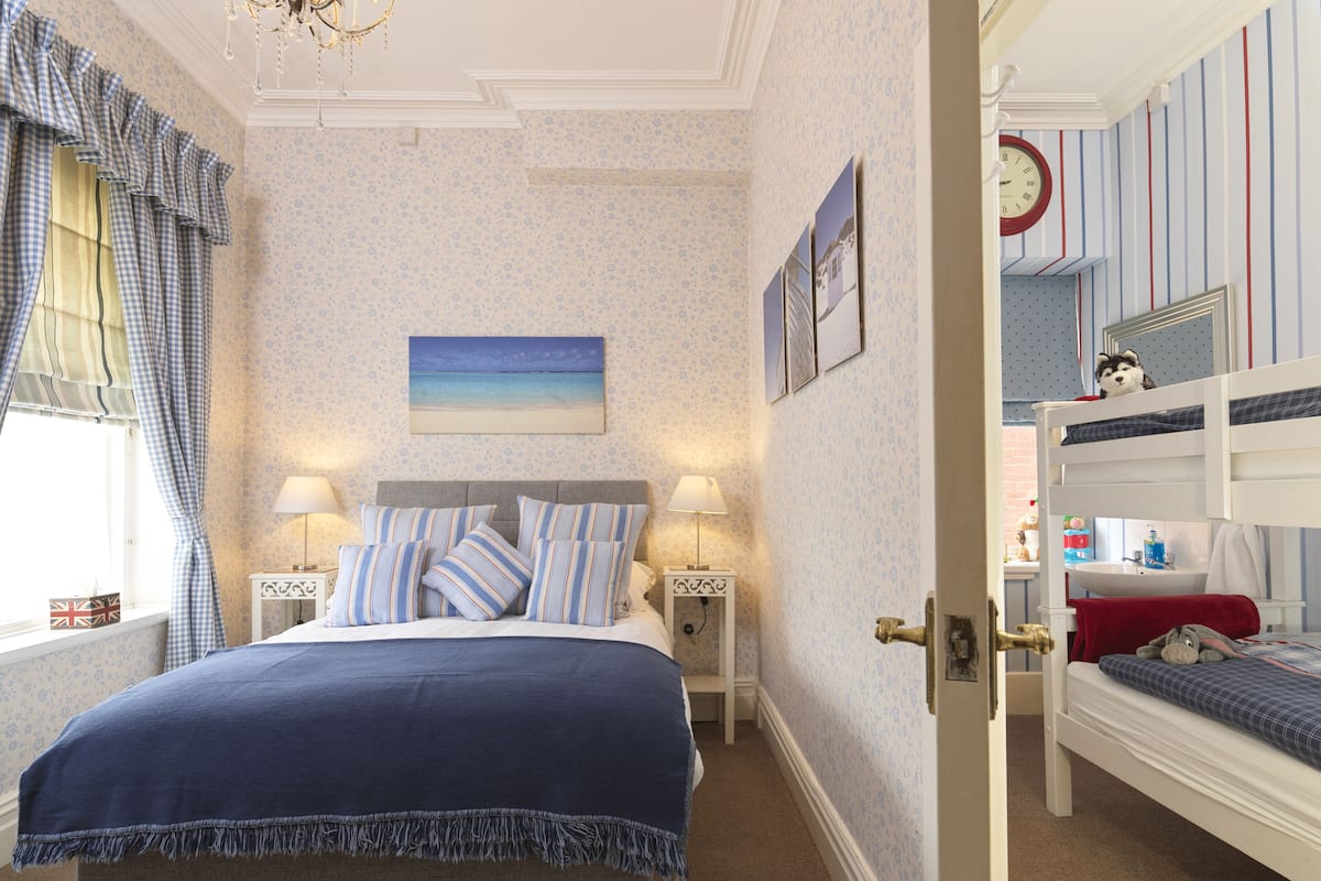 A bedroom with a bed, bedside tables, a nightstand, a dresser, a window with curtains, a wall clock, and a bunk bed with a stuffed animal.