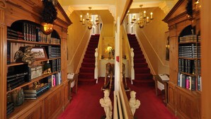 A grand hallway with a red carpet, wooden bookshelves filled with books, and a staircase with a chandelier. - Howarth House (Lytham St. Anne's)