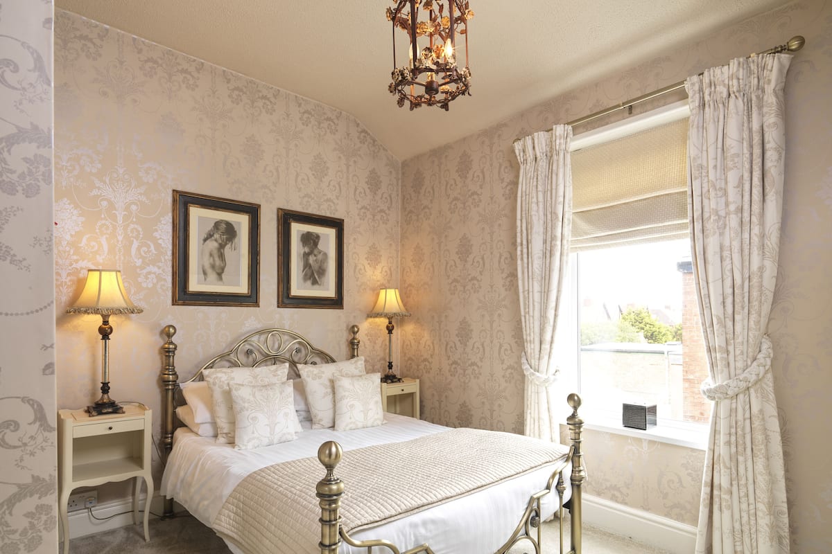 A classic bedroom with a four-poster bed, two bedside tables with lamps, and a large window with curtains.