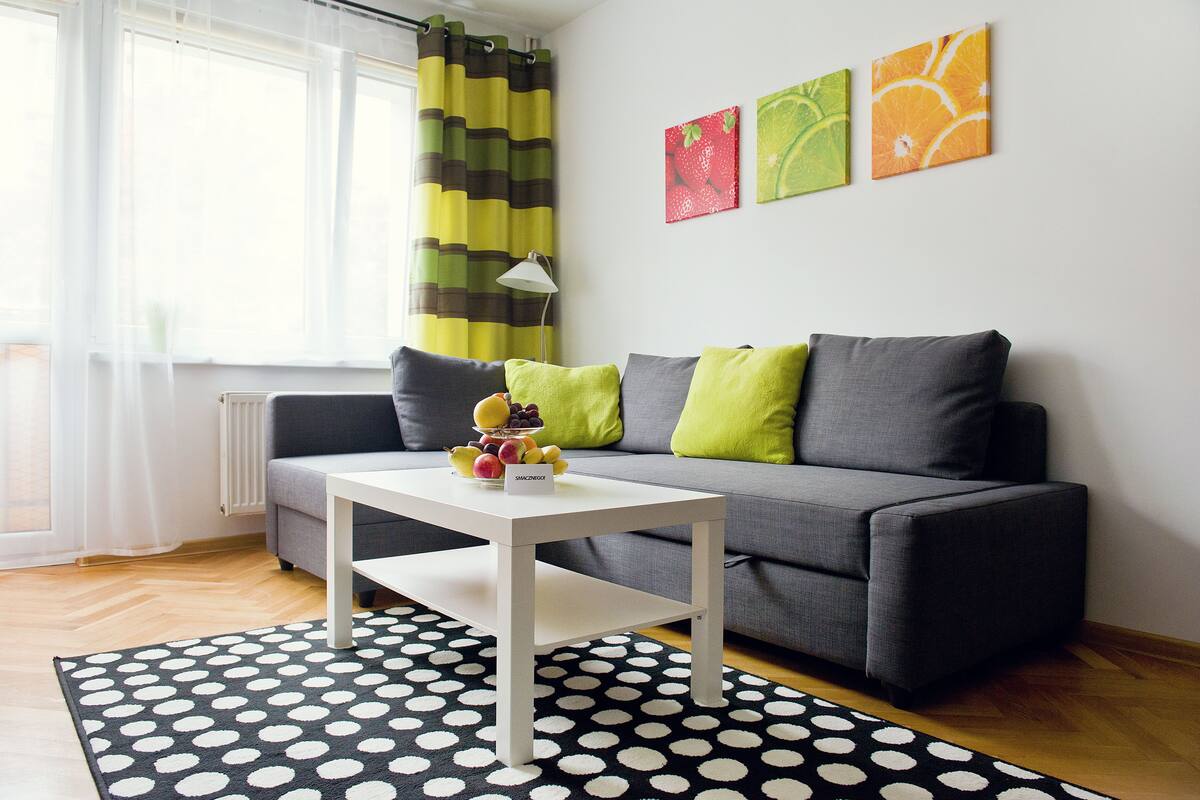 A modern living room with a grey sofa, a white coffee table, and a black and white polka dot rug. There are three fruit-themed wall art pieces and a striped curtain.