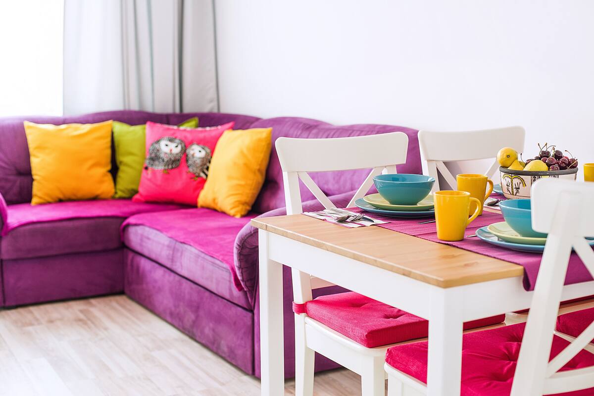 A cozy living room with a purple sofa, colorful cushions, a white chair, and a table set with yellow and blue dishes and cups.