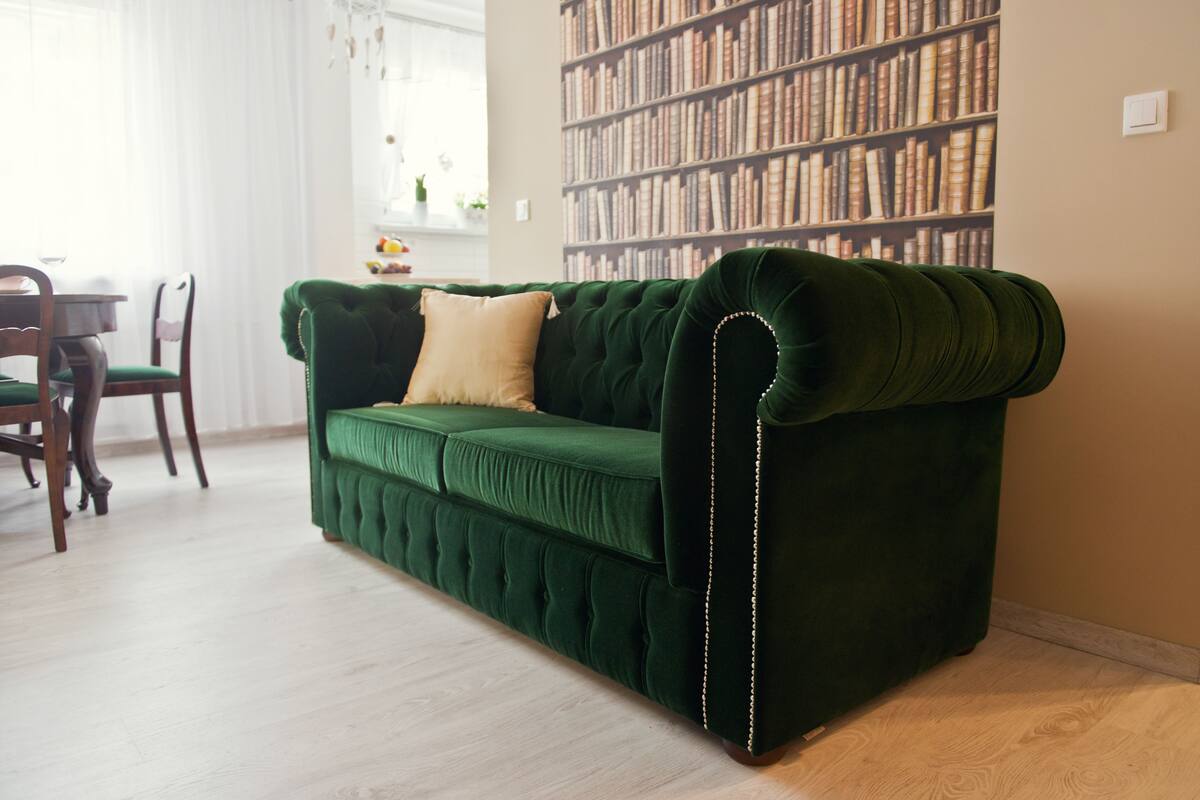 A green velvet sofa with a tufted backrest and a single beige pillow, positioned in a room with a wooden floor and a bookshelf filled with books in the background.
