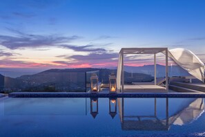 Una piscina infinita en la azotea con barandilla de cristal, dos faroles y vistas a montañas distantes al atardecer.
