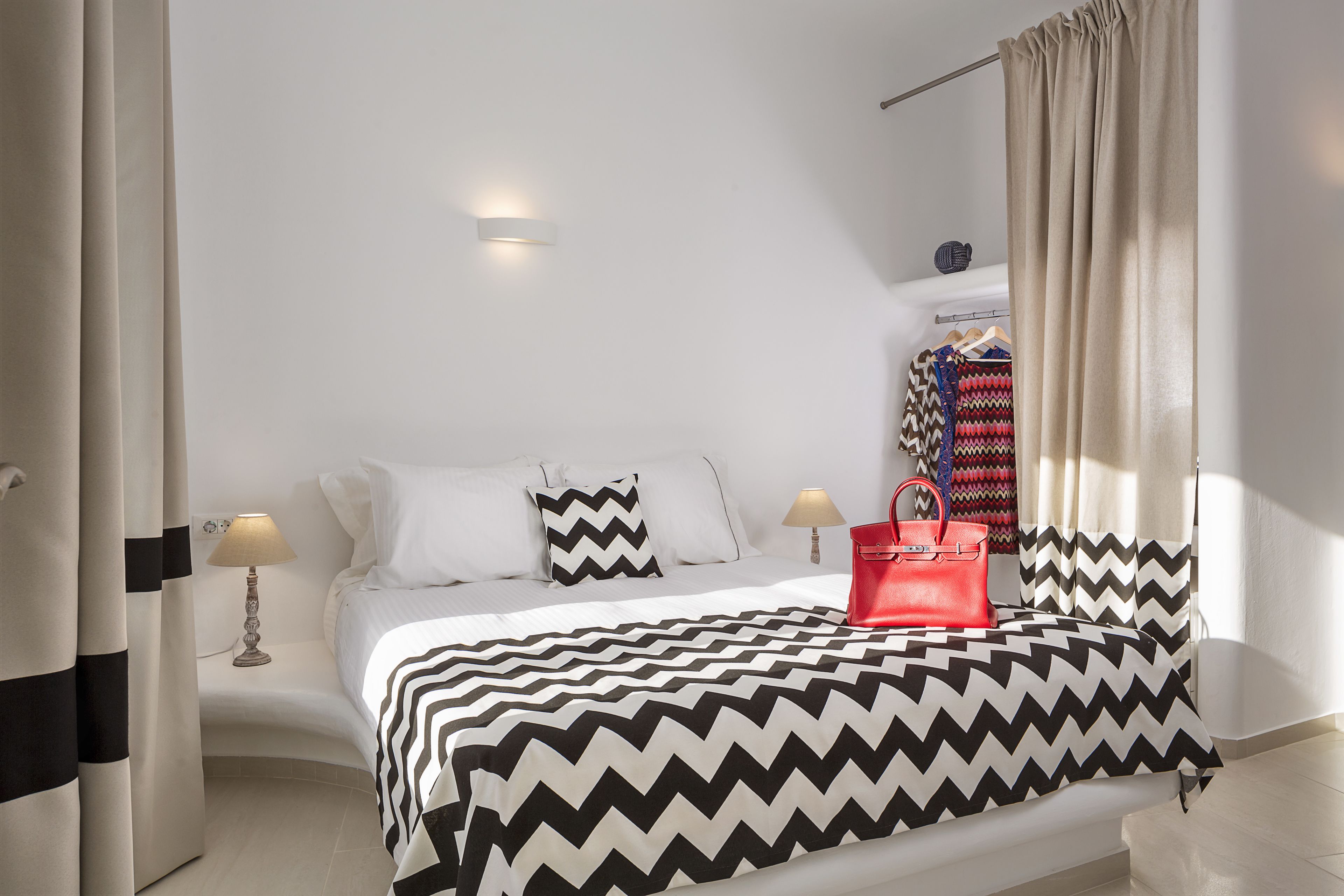 Ein Hotelzimmer mit einem Bett, zwei Nachttischlampen, einer roten Handtasche, einem Kleiderschrank und einem fenster mit Vorhängen.