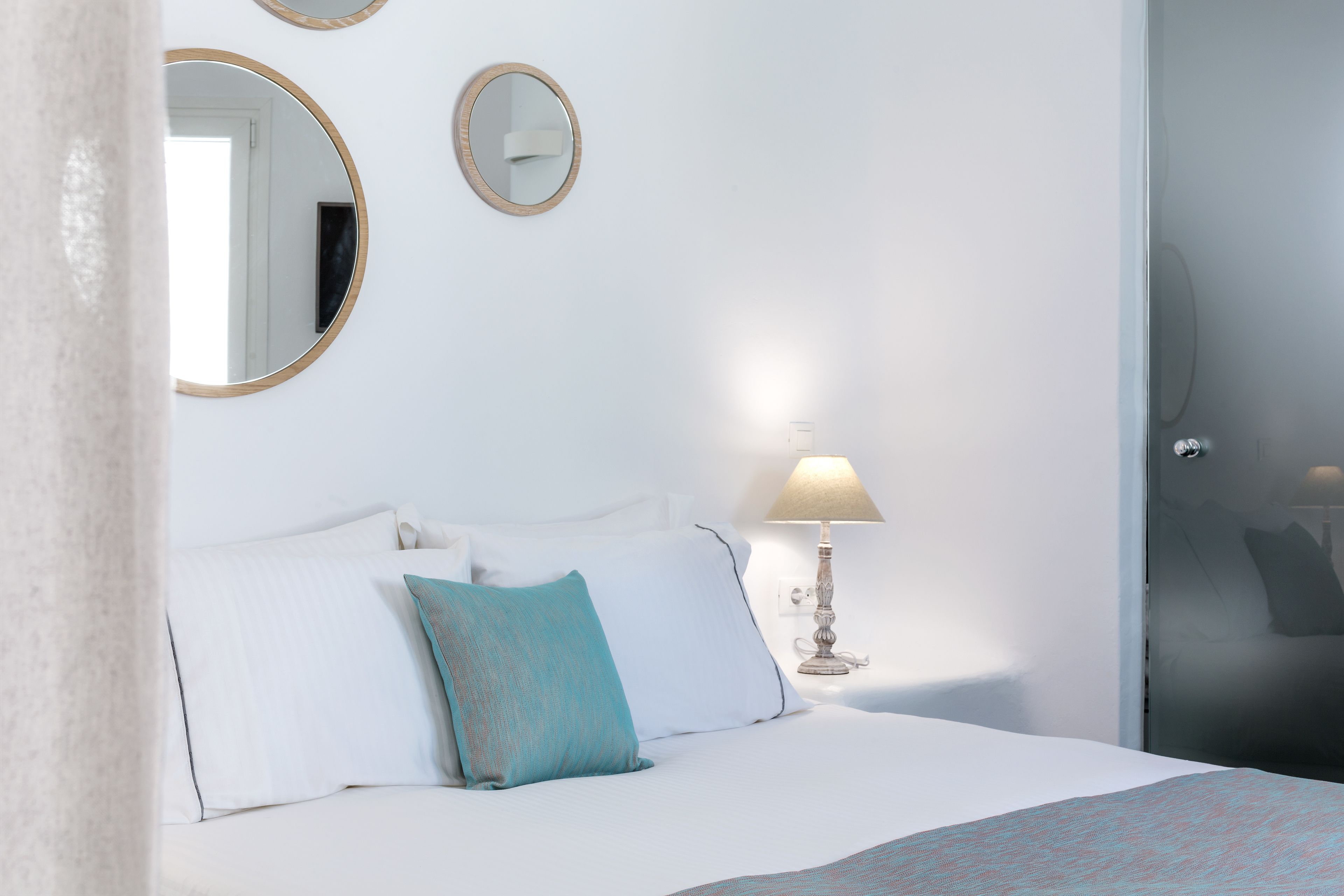 Une chambre d’hôtel avec un lit, du linge de lit blanc, un oreiller turquoise, une lampe et deux miroirs fixés au mur.