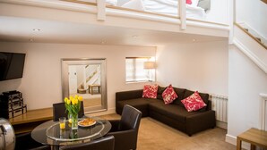 A modern living room with a sofa, a dining table with chairs, a TV, and a staircase leading to an upper level. - Howarth House Apartments (Lytham St. Anne's)