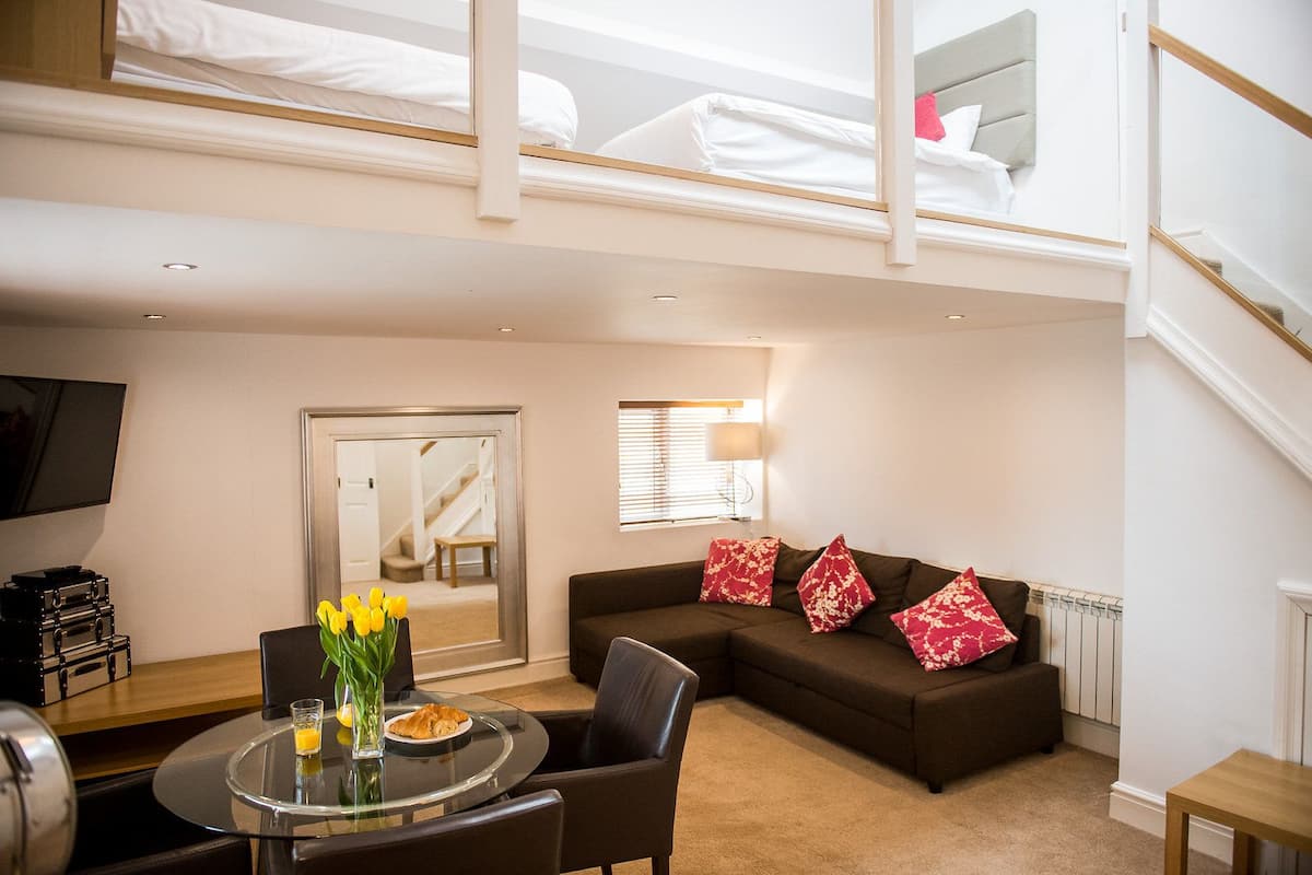 A modern living room with a sofa, a dining table with chairs, a TV, and a staircase leading to an upper level.
