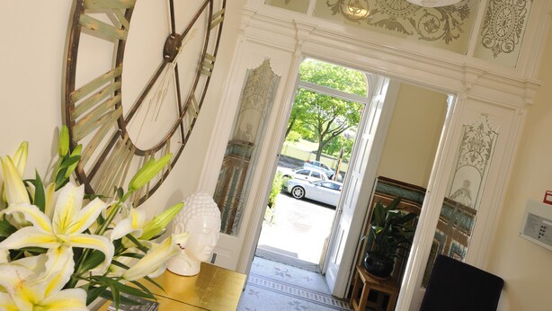 A room with a large clock, a vase of lilies, and a view of the street outside. - Howarth House Apartments (Lytham St. Anne's)