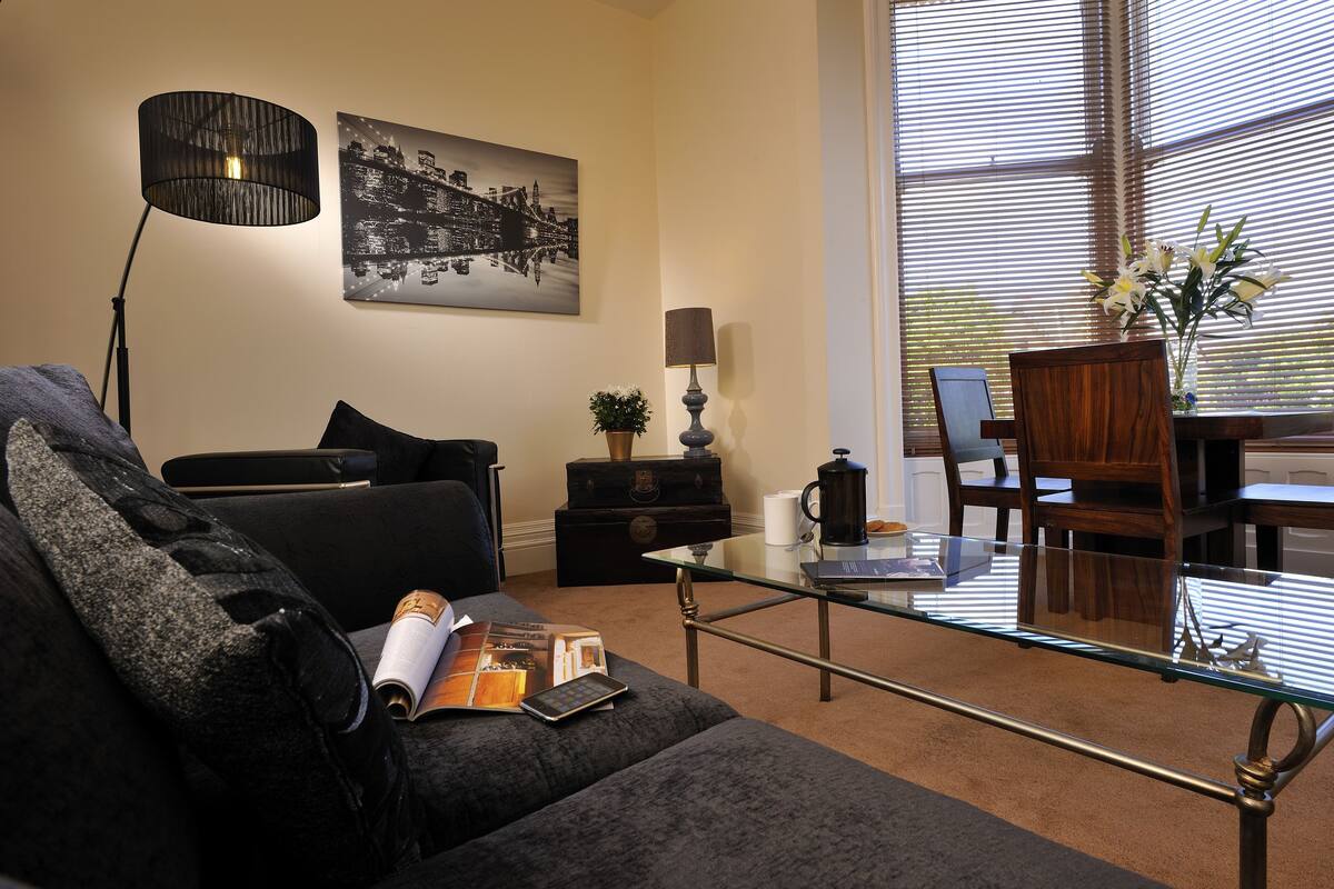 A modern living room with a glass coffee table, a black sofa, a lamp, a painting, and a vase with flowers.