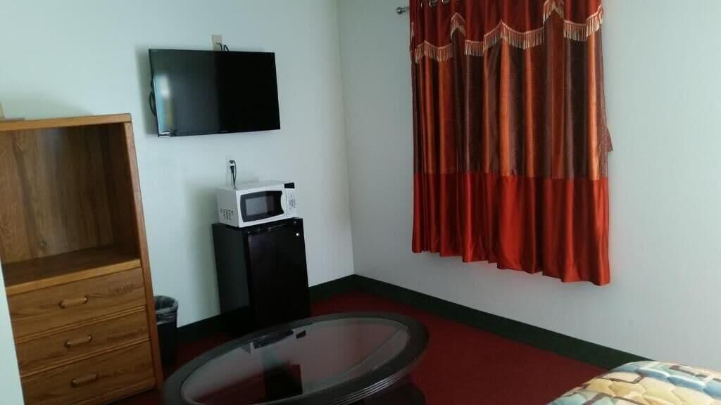 A room with a flat-screen TV mounted on the wall, a microwave on a small black cabinet, a wooden cabinet with drawers, a glass table, and a red curtain.