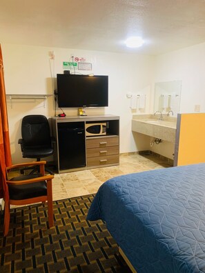 A hotel room with a bed, a desk with a television, a chair, a microwave, a sink, and a mirror.