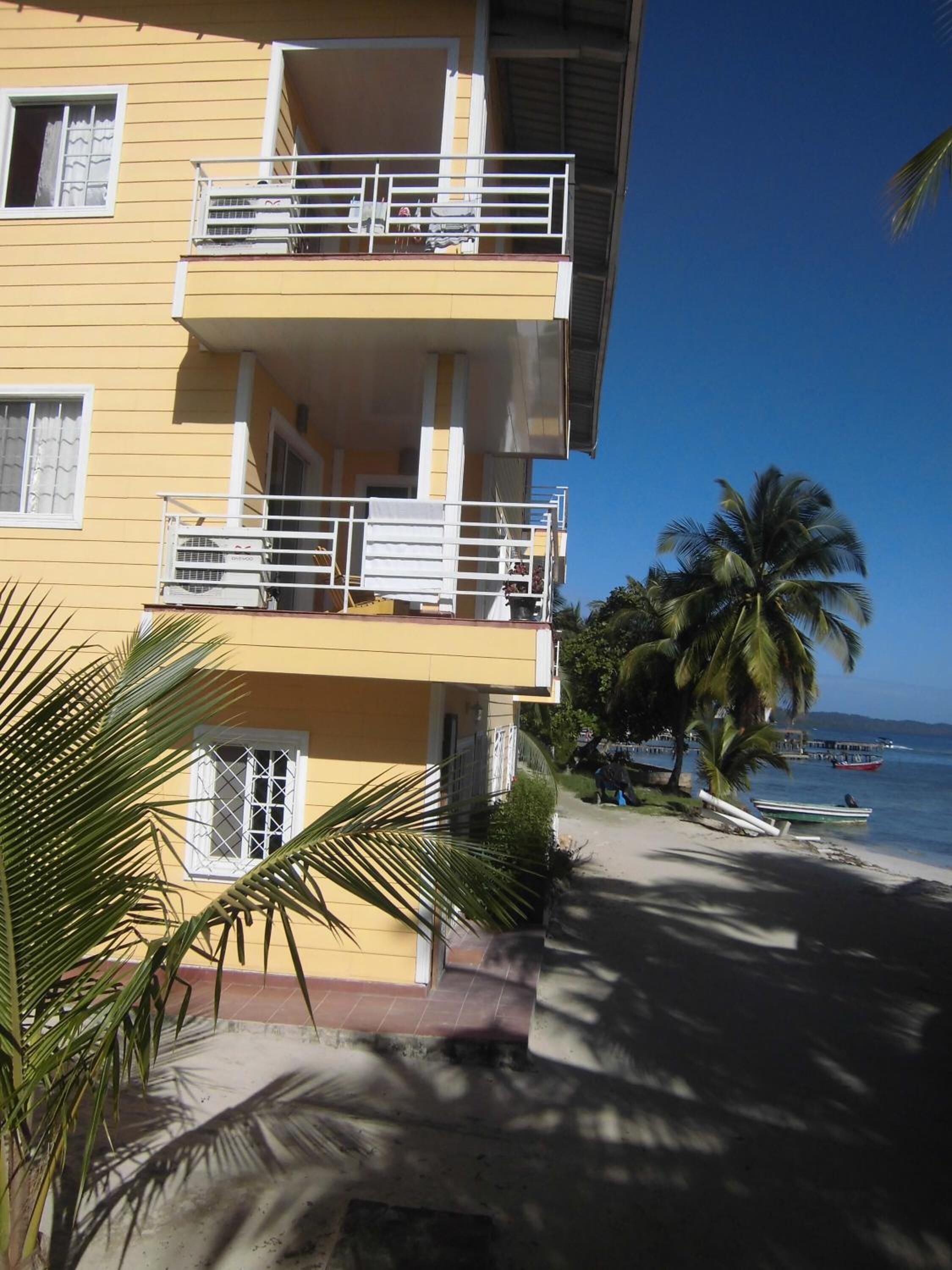 A yellow building with balconies, a palm tree, and a beach with boats in the background.