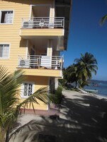 Un edificio amarillo con balcones, una palmera y una playa con barcos al fondo.