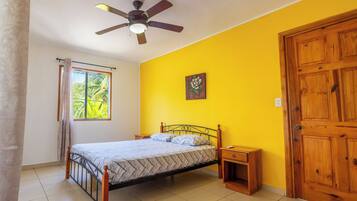 A bedroom with a bed, a ceiling fan, a window with curtains, a wooden door, and a small bedside table.