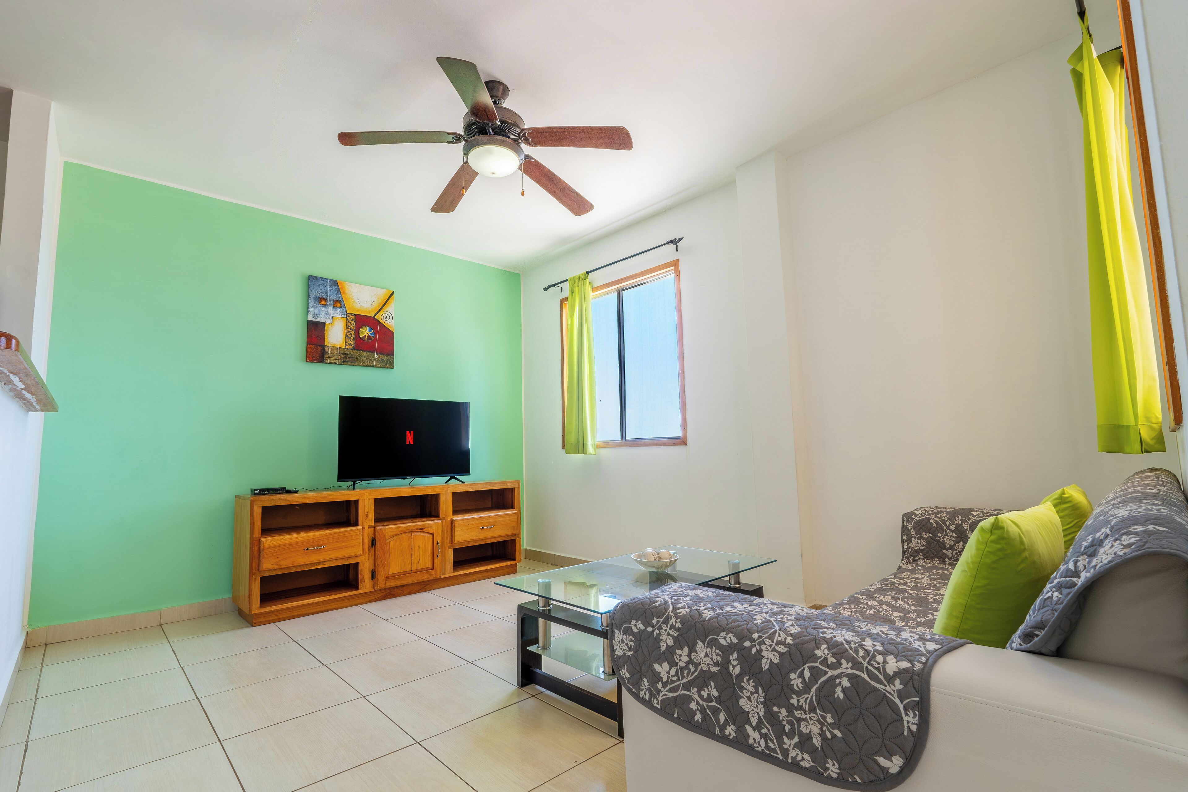 Una sala de estar con televisión, mueble de entretenimiento de madera, mesa de centro de vidrio, sofá y ventilador de techo.