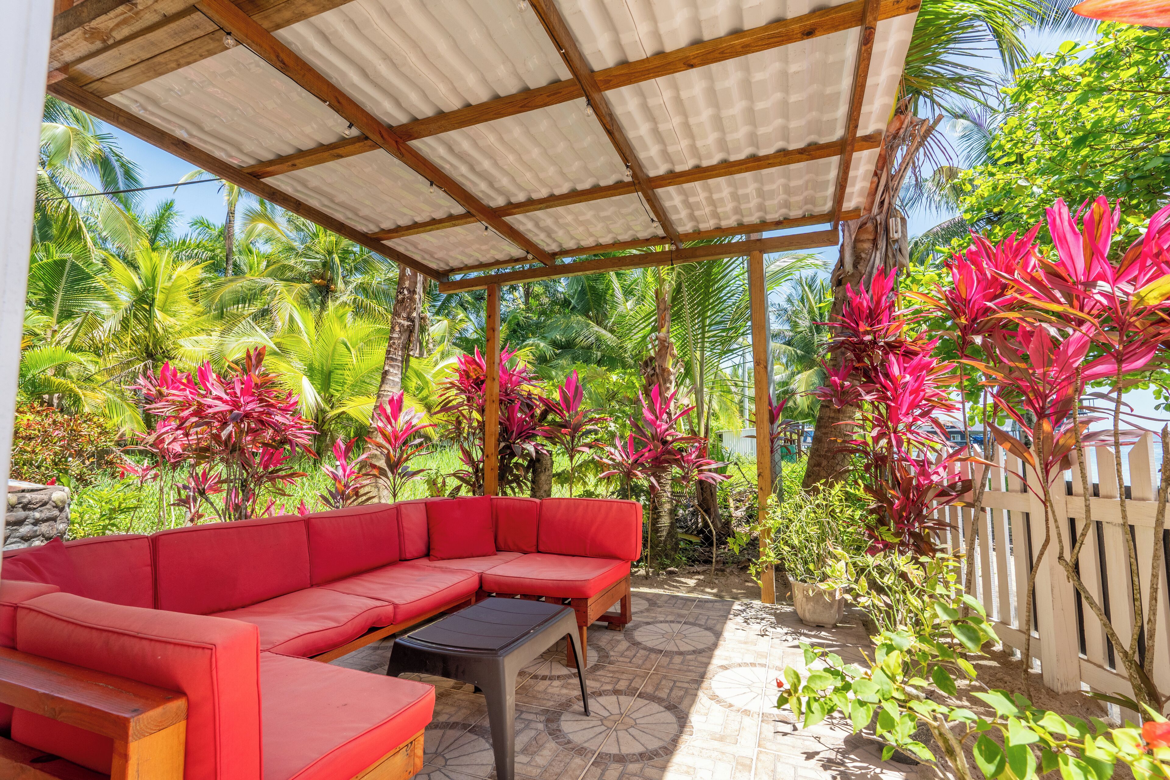 A covered outdoor seating area with a red sofa and a small table, surrounded by lush tropical plants and a wooden fence.