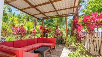 A covered outdoor seating area with a red sofa and a small table, surrounded by lush tropical plants and a wooden fence.