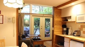A compact kitchen with wooden cabinets, a microwave, a coffee maker, and a toaster. There is a dining area with a table and chairs, and a small window with curtains. - Sea Star Beach Retreat (Tofino)