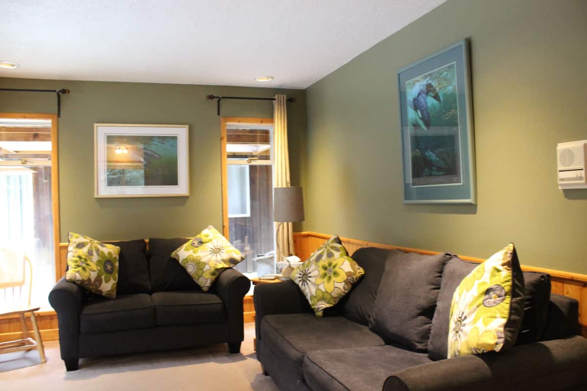 A living room with two sofas, a table, and a lamp. There are framed pictures on the wall.