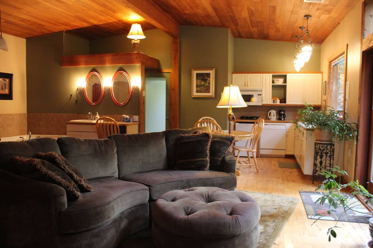 A cozy living room with a sectional sofa and ottoman, a kitchen in the background, wooden ceiling, and pendant lighting.