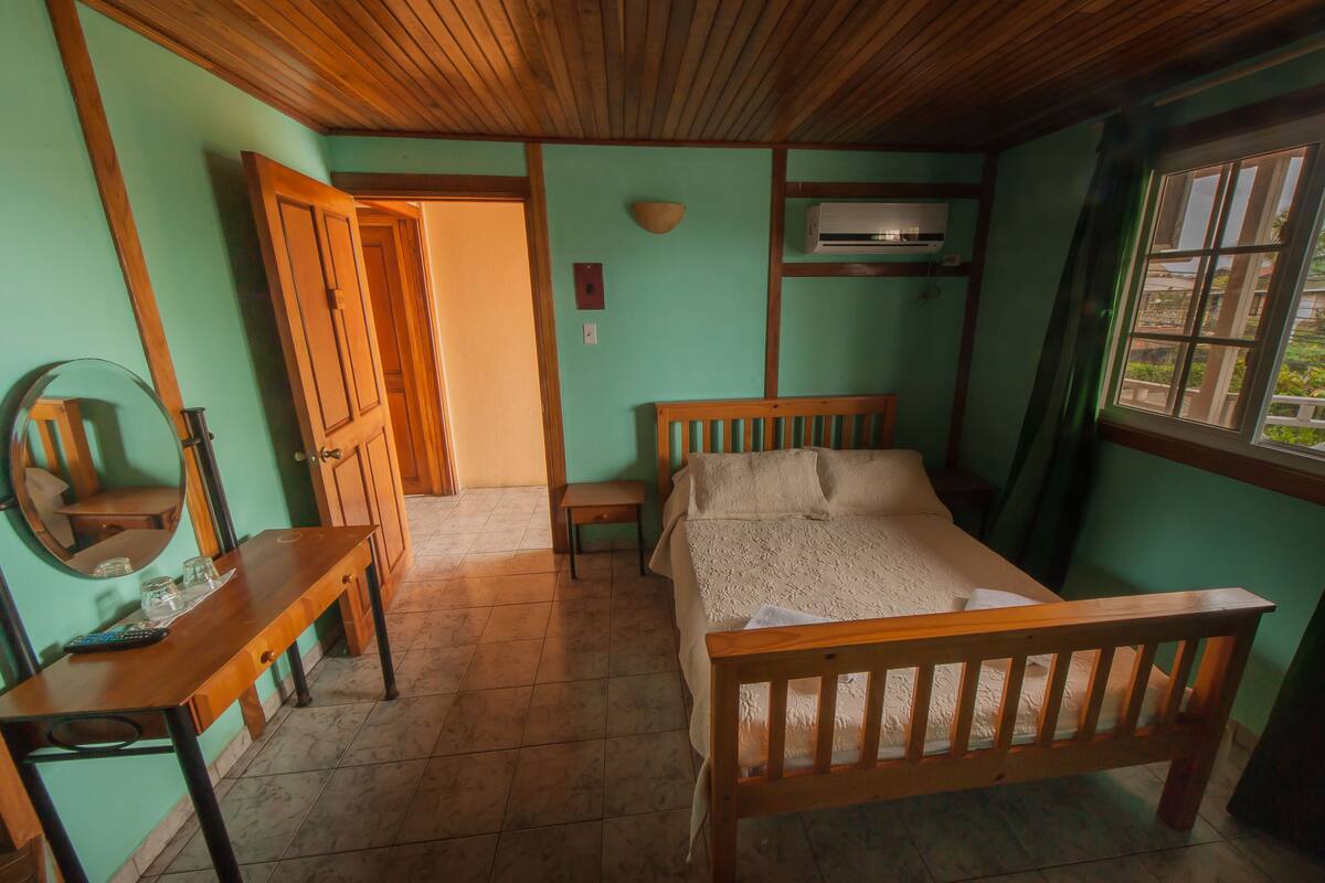 A room with a wooden bed, a wooden desk with a mirror, a small table, and a window with a view.