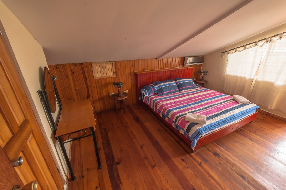 A bedroom with wooden walls, a bed with striped bedding, a wooden desk, and a small table with a lamp.