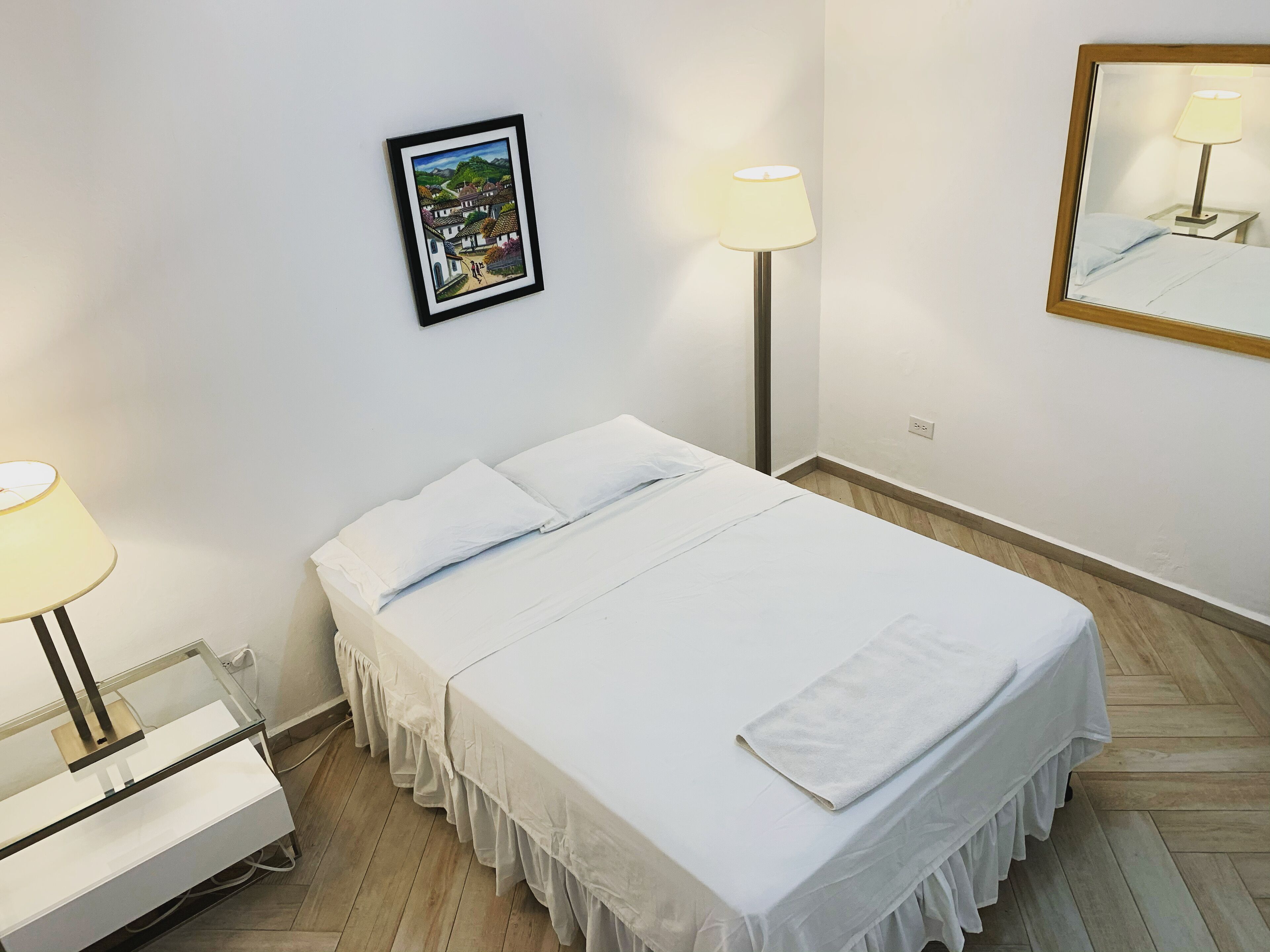A neatly made bed with white linens, a bedside table with a lamp, a framed picture on the wall, and a mirror reflecting the lamp.